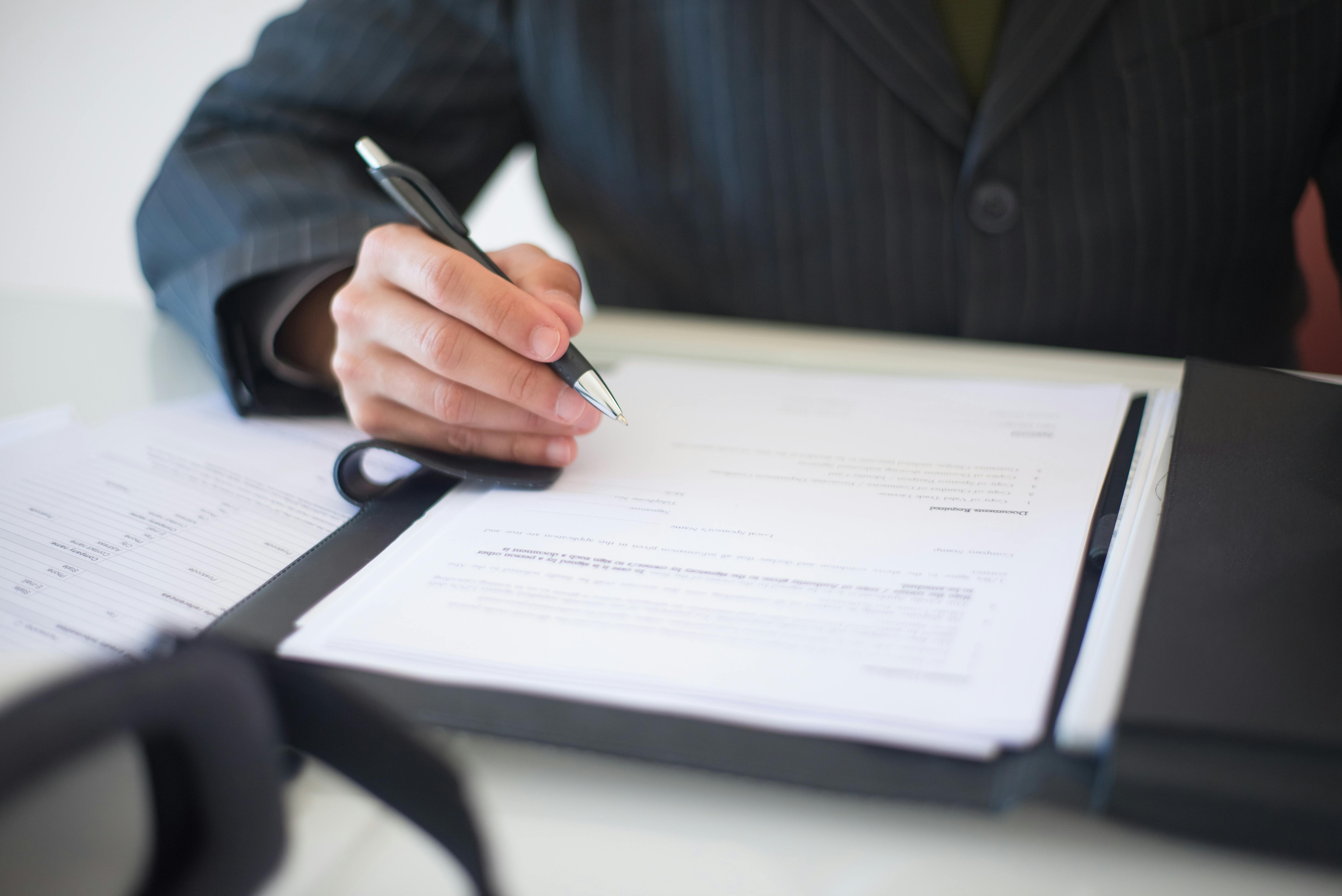 A person sits at a table with an open folder in front of them, and a white document inside the folder. They hold a pen for signing the document.